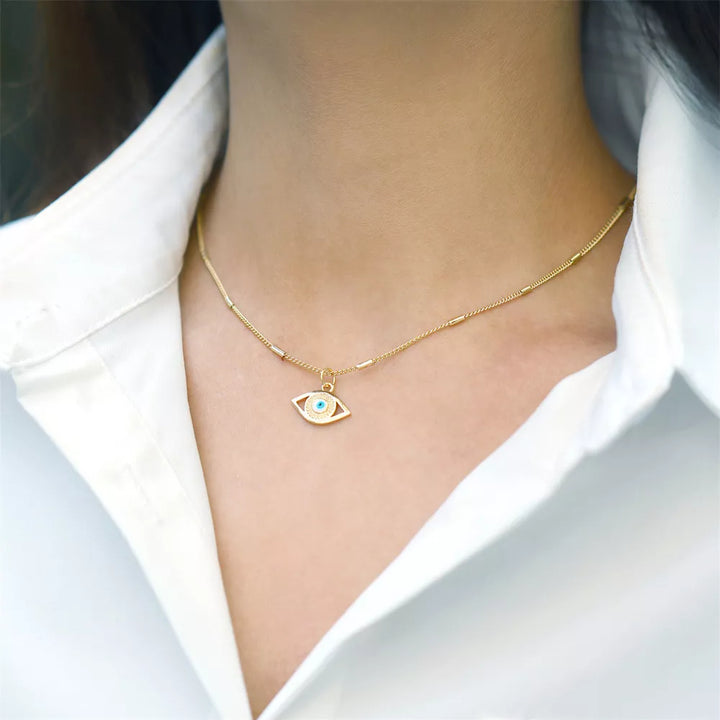 A woman wears a white collared shirt and a gold evil eye necklace featuring a blue zirconia stone at its center, symbolizing protection. The necklace is hypoallergenic, making it comfortable for everyday wear.