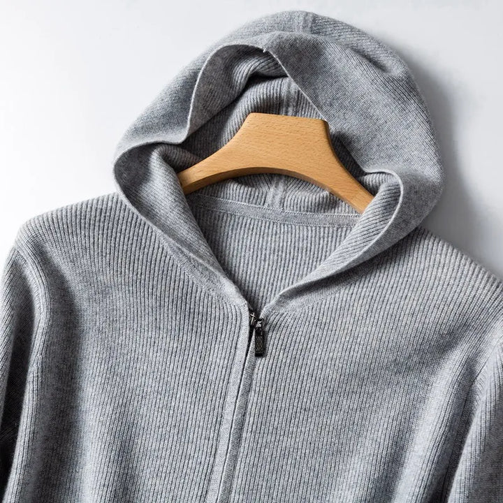 A close-up of a light gray, ribbed cashmere hoodie hanging on a wooden hanger against a white background. The hood is spread open, revealing the lightweight insulating texture of the fabric.
