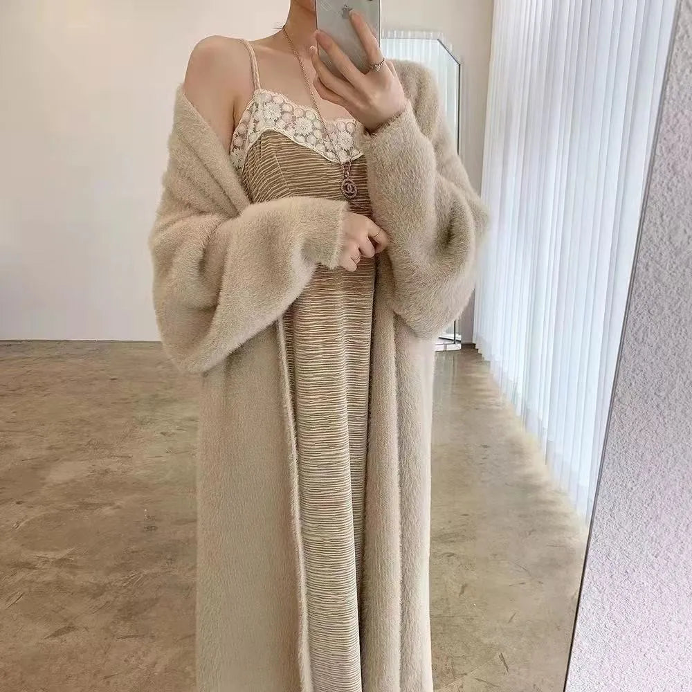 A person takes a mirror selfie wearing a long, beige cardigan with a relaxed drape fit over a light-colored, lace-trimmed dress. The face is not visible, and the setting is a minimalistic room with light walls and floor.