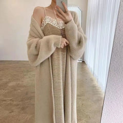 A person takes a mirror selfie wearing a long, beige cardigan with a relaxed drape fit over a light-colored, lace-trimmed dress. The face is not visible, and the setting is a minimalistic room with light walls and floor.