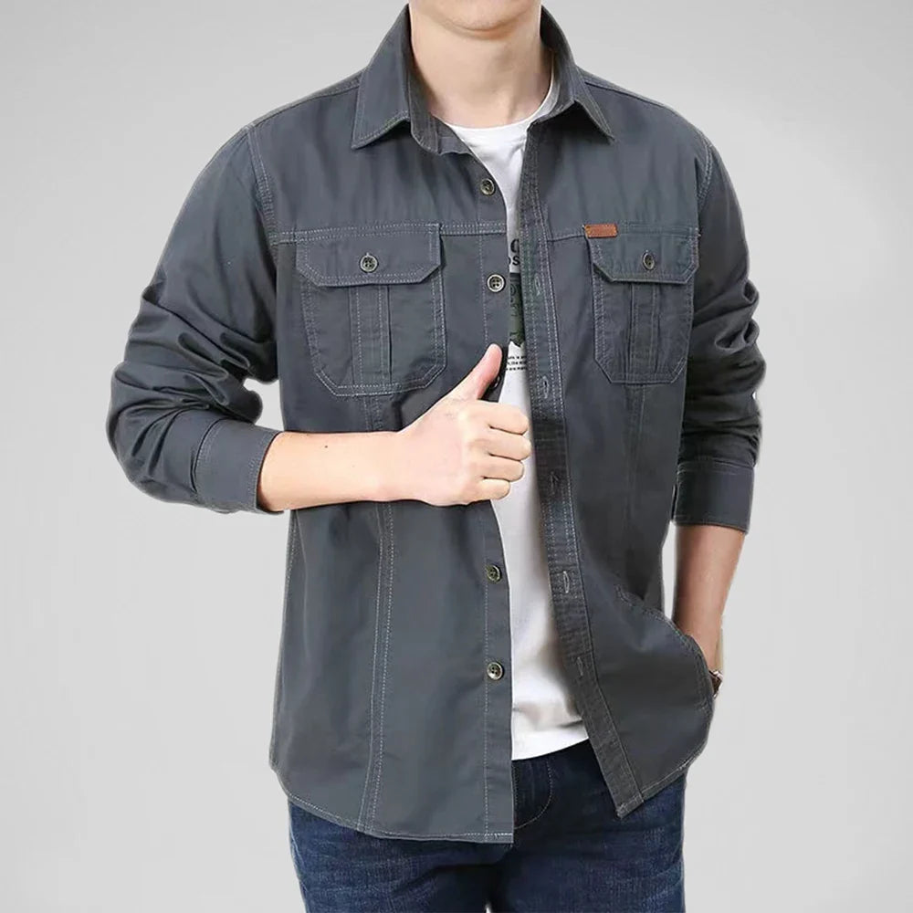 A person wearing a dark gray relaxed fit button-up shirt over a breathable undershirt and jeans stands with one hand in their pocket and the other giving a thumbs up against a plain gray background.