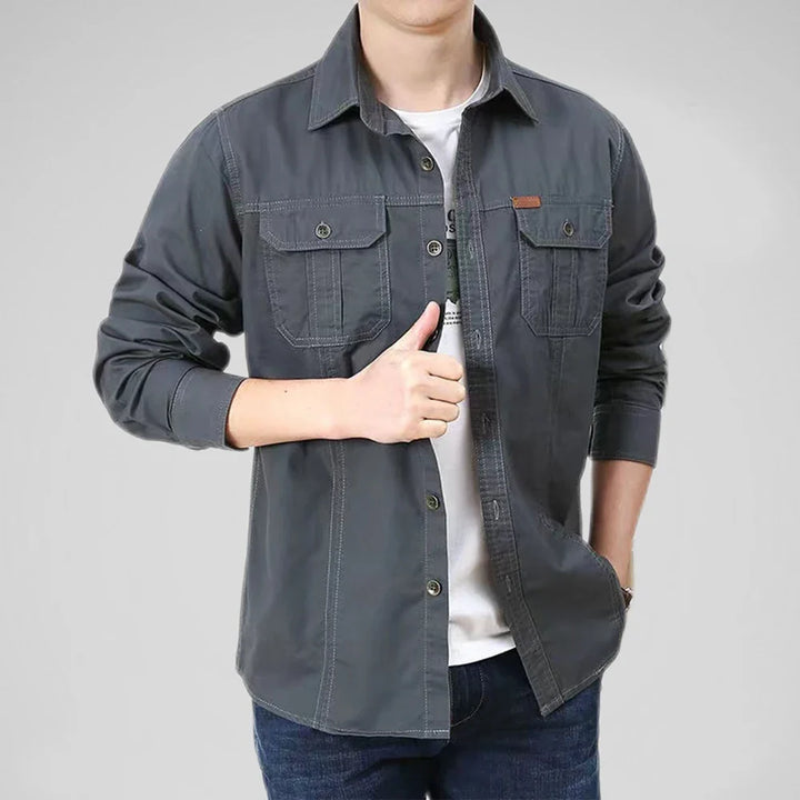 A person wearing a dark gray relaxed fit button-up shirt over a breathable undershirt and jeans stands with one hand in their pocket and the other giving a thumbs up against a plain gray background.