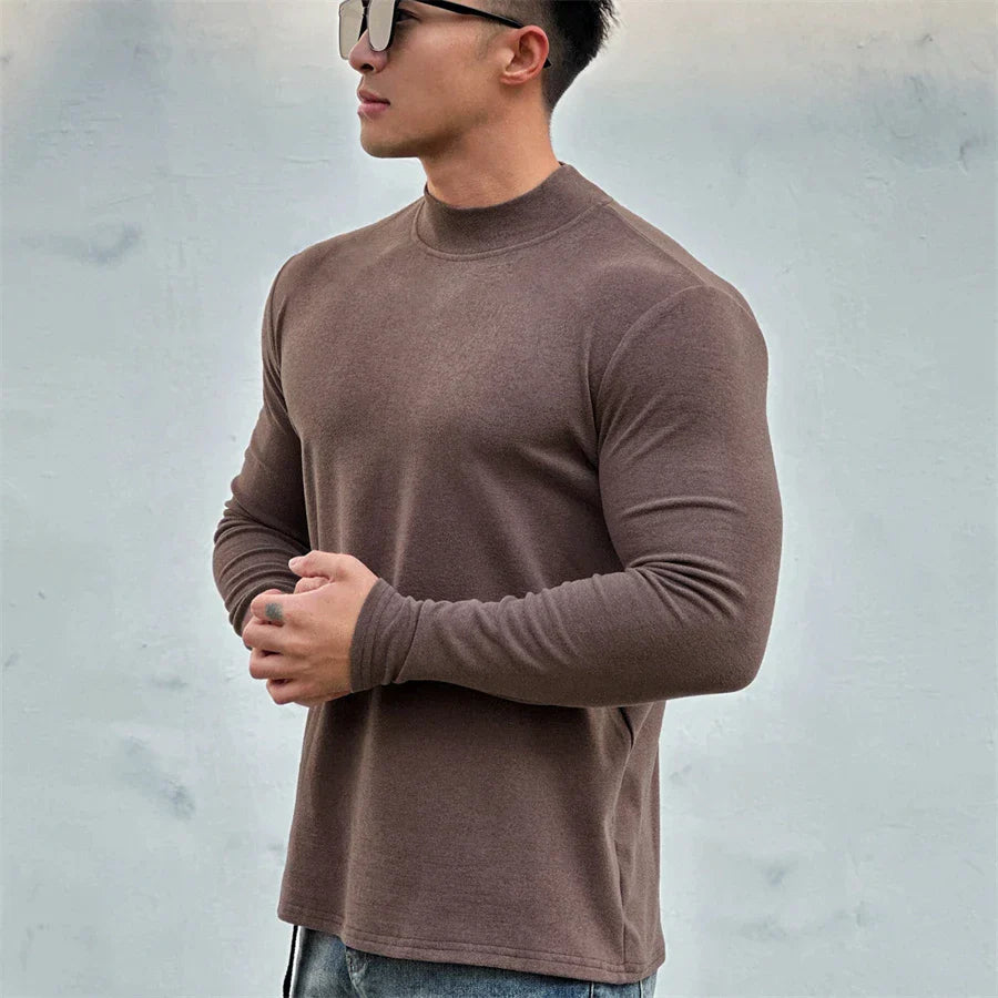 A man wearing sunglasses and a close-fitting brown long-sleeve shirt with a mock neck collar stands against a plain light gray background, holding his hands together in front of him.