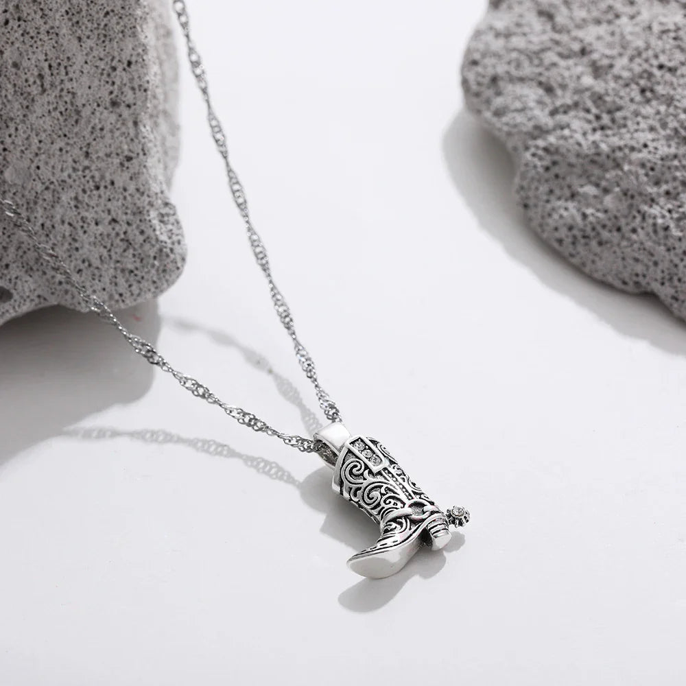 A silver cowgirl necklace with a pendant shaped like a detailed cowboy boot rests on a white surface between two light gray textured stones.