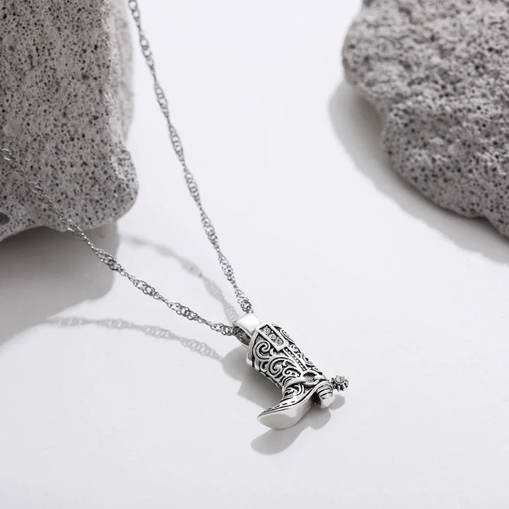 A silver cowgirl necklace with a pendant shaped like a detailed cowboy boot rests on a white surface between two light gray textured stones.