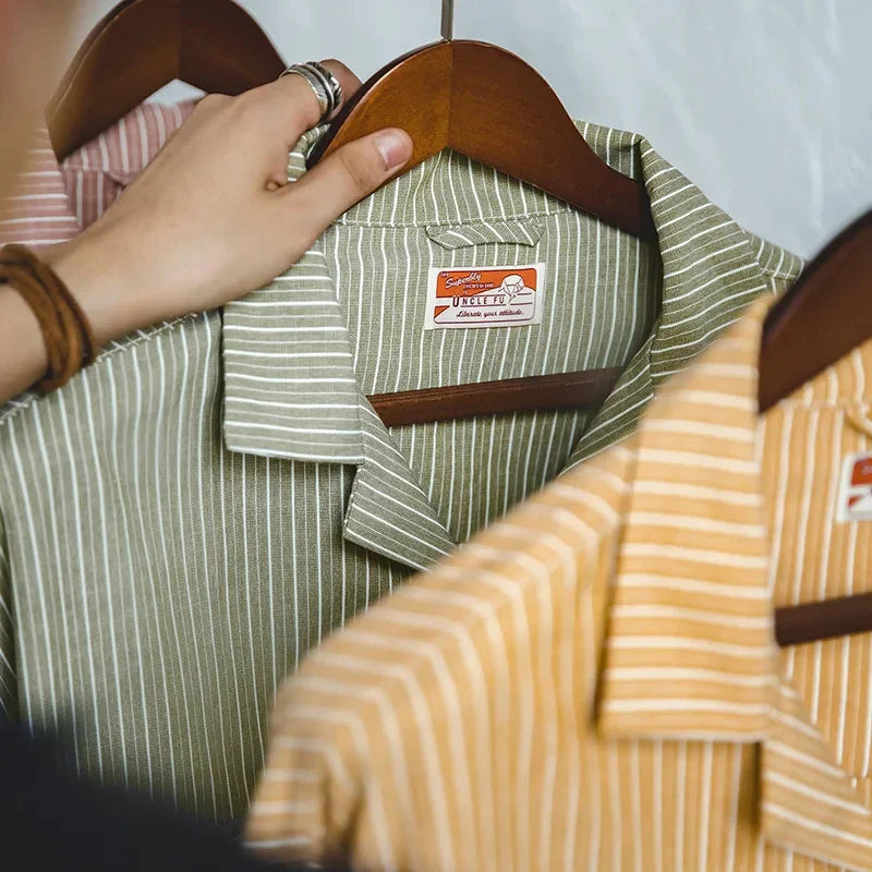 A hand holds a wooden hanger with a green and white Cuban collar shirt. Next to it are similar high-quality cotton shirts in yellow and orange stripes, all hanging neatly on wooden hangers.