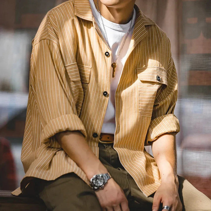 A person wearing a tan and white pinstripe shirt over a white t-shirt, olive pants, a brown belt, watch, and necklace sits with arms resting on their knees. The high-quality cotton fabric adds comfort; face is partially out of frame.