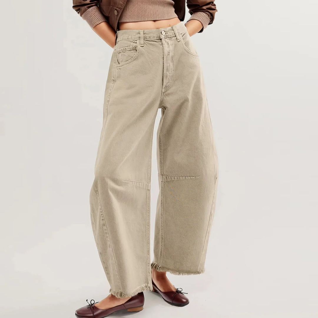 A person wearing relaxed fit, high-waisted beige jeans with frayed hems, a cropped brown sweater, and brown ballet flats stands against a plain light background. The image focuses on the pants and lower body.