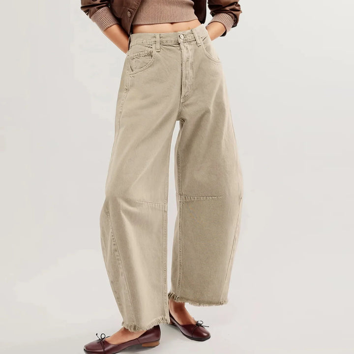 A person wearing relaxed fit, high-waisted beige jeans with frayed hems, a cropped brown sweater, and brown ballet flats stands against a plain light background. The image focuses on the pants and lower body.