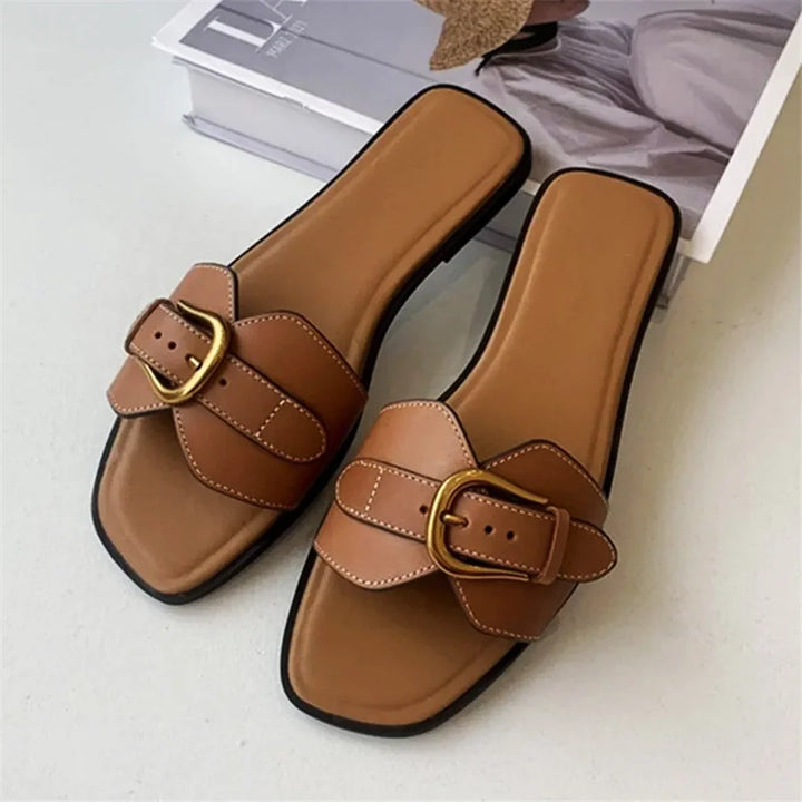 A pair of brown leather slides with wide straps and gold buckles are placed on a light surface next to a magazine. These slip-on sandals feature square-shaped toes and black soles.