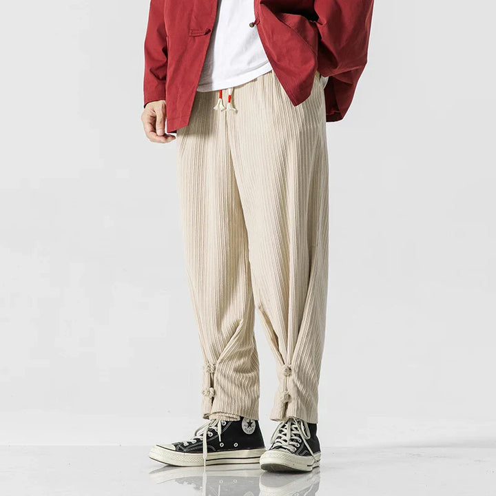 A person wearing beige cotton blend sweatpants with a drawstring waistband and ties at the ankles, a white shirt, a red jacket, and black high-top sneakers stands against a plain light background.