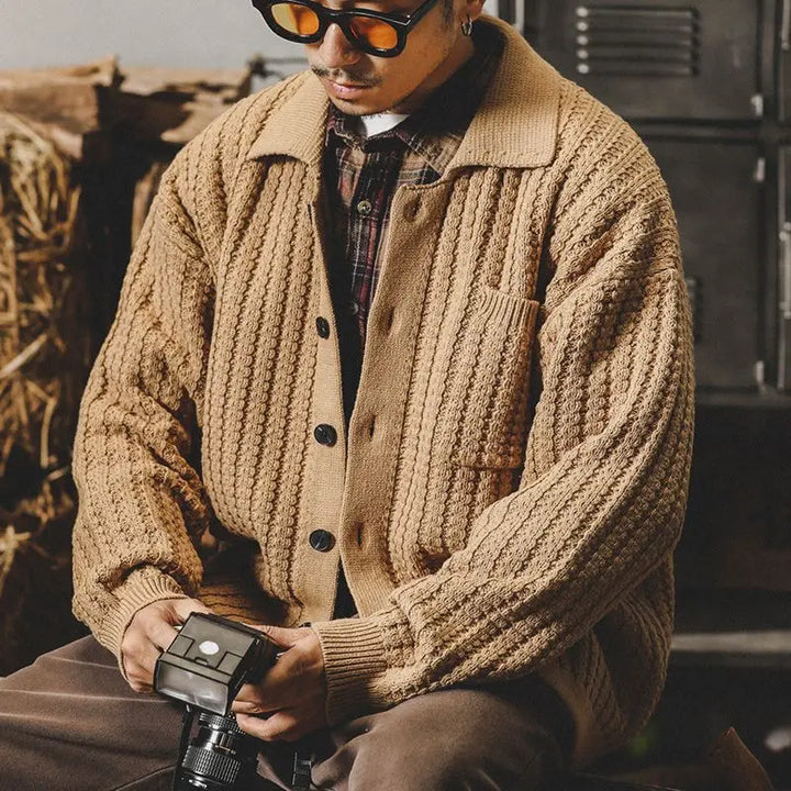 A man in a wool cardigan with a heritage aesthetic sits indoors, gazing down as he holds a vintage camera. He sports sunglasses and a cap, with straw bales and dark lockers providing the backdrop.