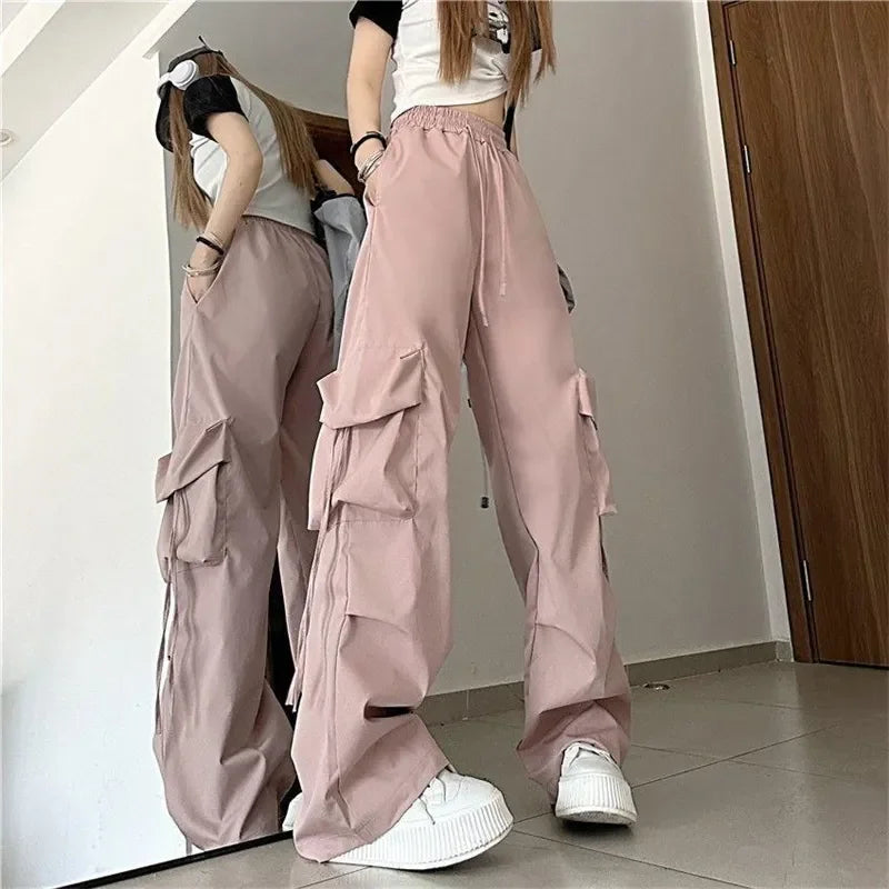 A person wearing loose, light pink cargo pants with a high-waist and multiple cargo pockets, a white t-shirt with black sleeves, and white sneakers stands indoors in front of a mirror, partially reflected.