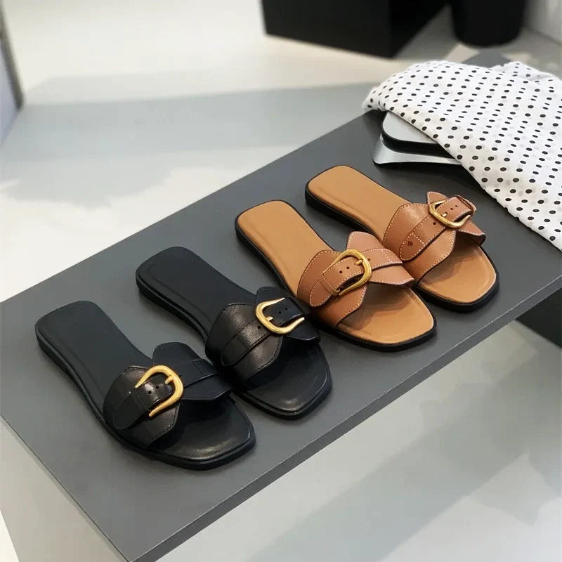 Two pairs of leather slides—one black and one tan, each featuring a gold buckle—are displayed on a gray table next to a white polka-dotted fabric.