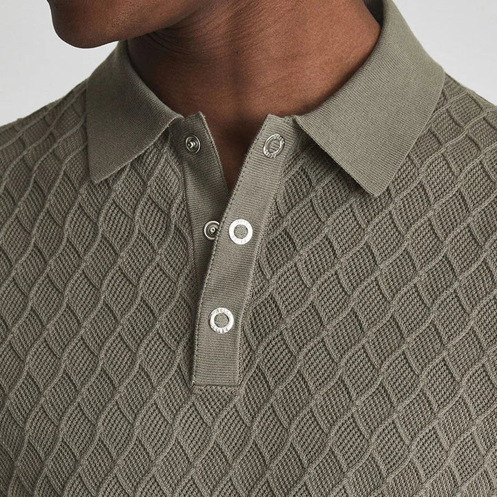 Close-up of a person wearing a wrinkle-resistant, olive-green herringbone polo with a diamond pattern and three white buttons on the placket. The image shows the shirt collar and upper chest area.