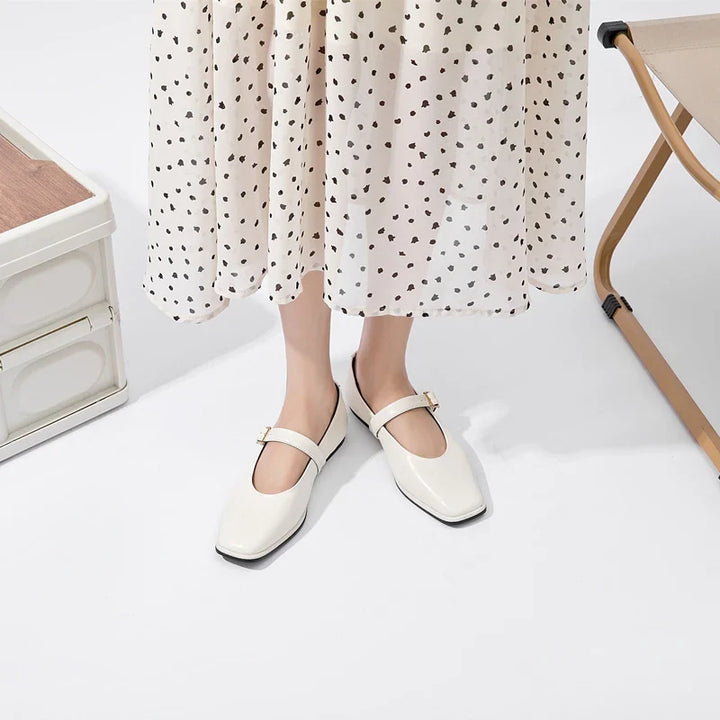 A person wearing a white, polka dot dress and cream-colored handmade leather flats stands on a white floor next to a beige folding chair and a white cabinet.