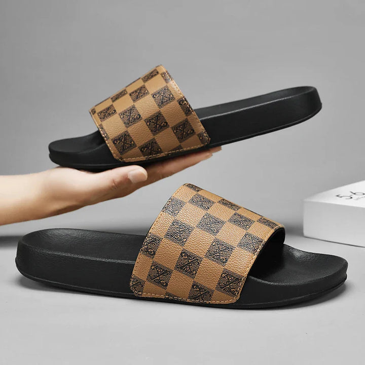 A hand holds a black streetwear slide with a wide tan and brown checkerboard strap featuring a subtle decorative pattern. Another identical checkered sandal lies flat on a gray surface.