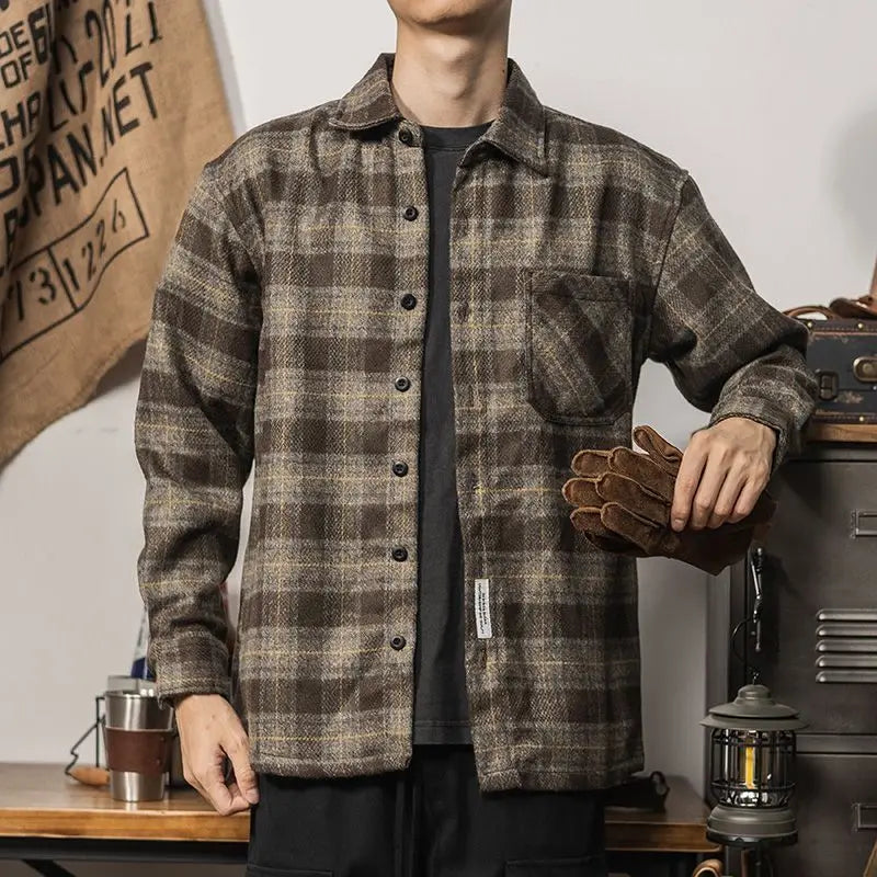 A person wearing a relaxed fit brown plaid button-up shirt stands indoors, holding brown work gloves in one hand. The background includes a hanging burlap sack and camping gear like a lantern and metal cups.