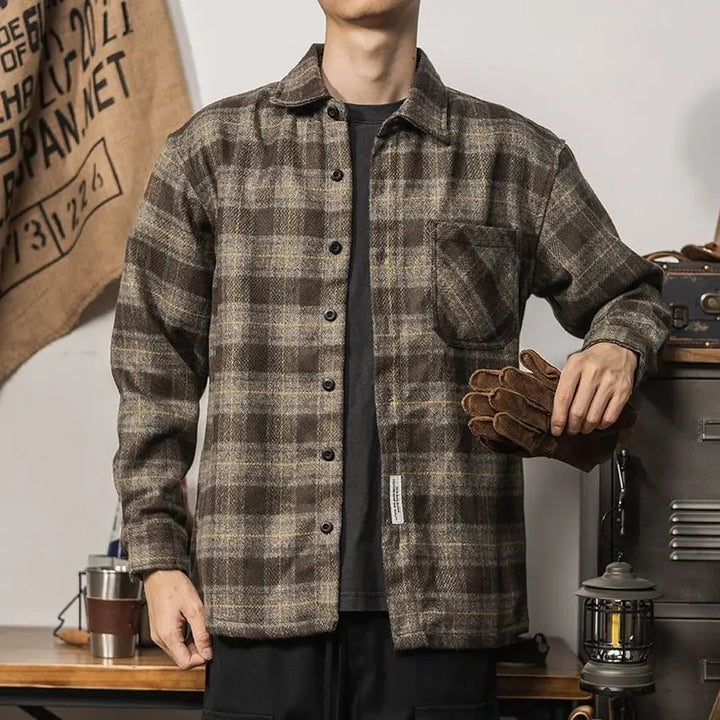 A person wearing a relaxed fit brown plaid button-up shirt stands indoors, holding brown work gloves in one hand. The background includes a hanging burlap sack and camping gear like a lantern and metal cups.