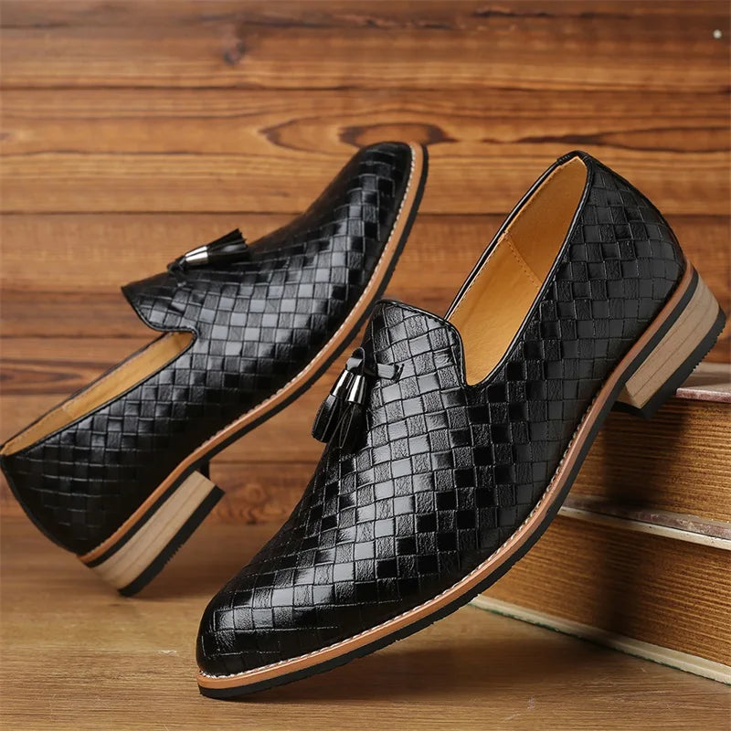 A pair of black, woven leather loafers with tassels and a classic basketweave pattern, featuring tan soles and low heels, displayed against a wooden background.