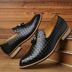 A pair of black, woven leather loafers with tassels and a classic basketweave pattern, featuring tan soles and low heels, displayed against a wooden background.