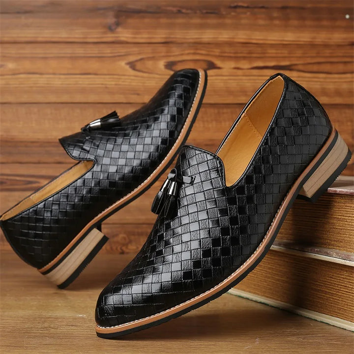 A pair of black, woven leather loafers with tassels and a classic basketweave pattern, featuring tan soles and low heels, displayed against a wooden background.