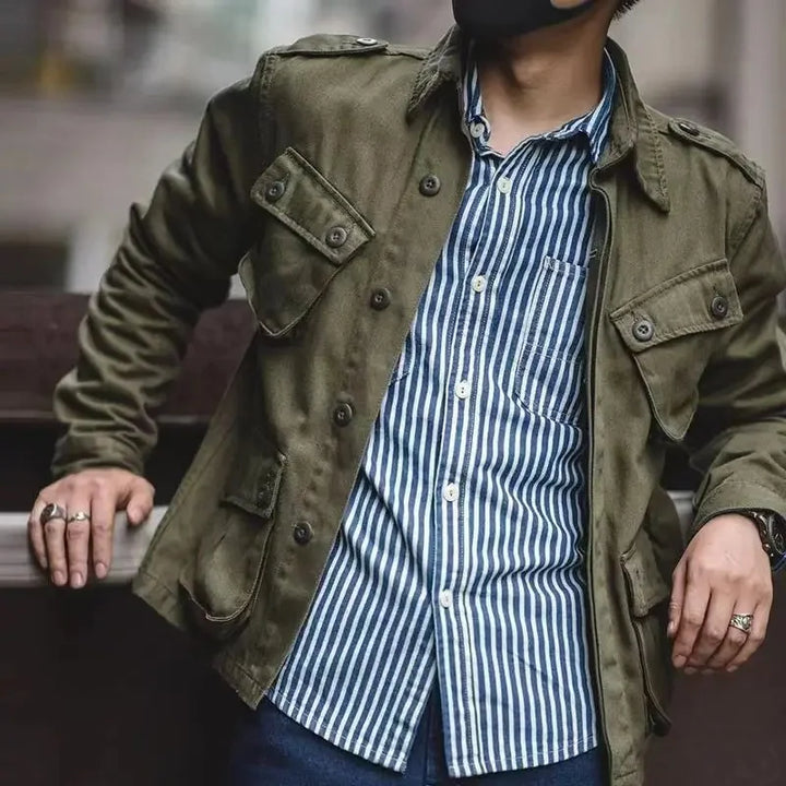 A person wearing a vintage military jacket over a blue and white vertically striped button-up shirt, dark jeans, and several rings, leans against a railing with their face partially out of frame.