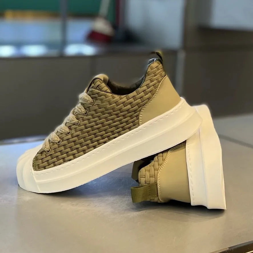 A pair of beige woven upper sneakers with thick white cushioned soles is displayed on a smooth surface; one shoe stands upright while the other is laid on its side beneath it.