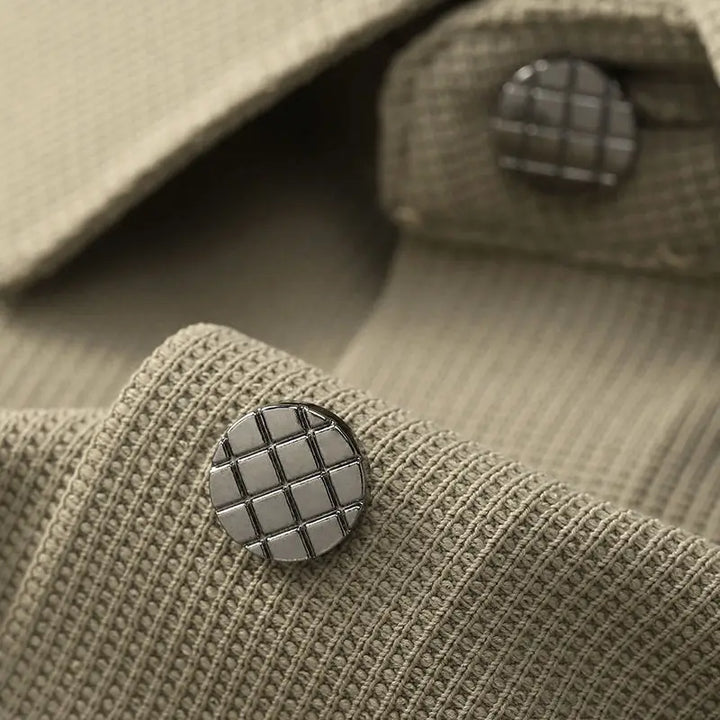 Close-up of a textured beige fabric from an anti-wrinkle dress shirt, with a metallic button featuring a grid pattern. Another similar button is visible in the background.