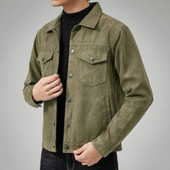 A person wearing an olive green button-up suede jacket with a classic point collar over a black turtleneck, one hand touching the jacket and the other in their pocket, photographed against a plain gray background.