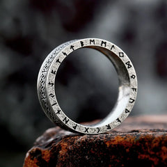 A durable metal ring engraved with runic symbols and intricate patterns rests upright on a rough, dark stone surface, its design reminiscent of an ancient rune ring against the blurred background.