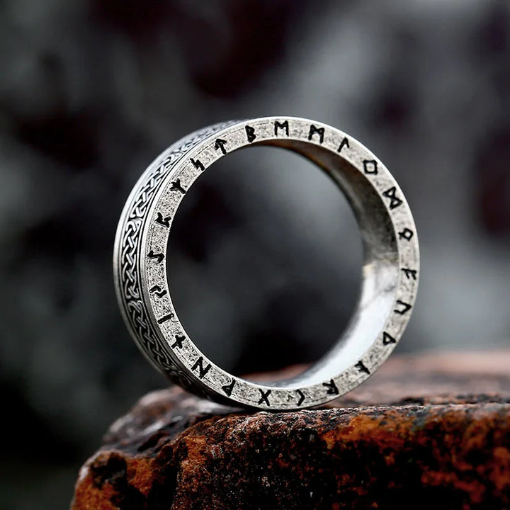 A durable metal ring engraved with runic symbols and intricate patterns rests upright on a rough, dark stone surface, its design reminiscent of an ancient rune ring against the blurred background.