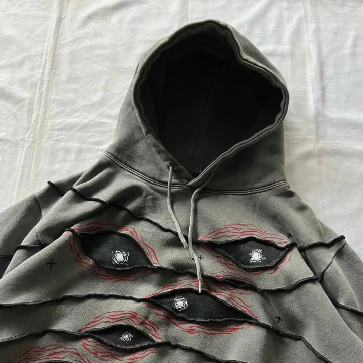 A gray hoodie with a comfortable loose fit features layered, jagged fabric and an eye pattern patchwork outlined in red and black, giving a textured, surreal look. The hoodie is laid flat on a white surface.