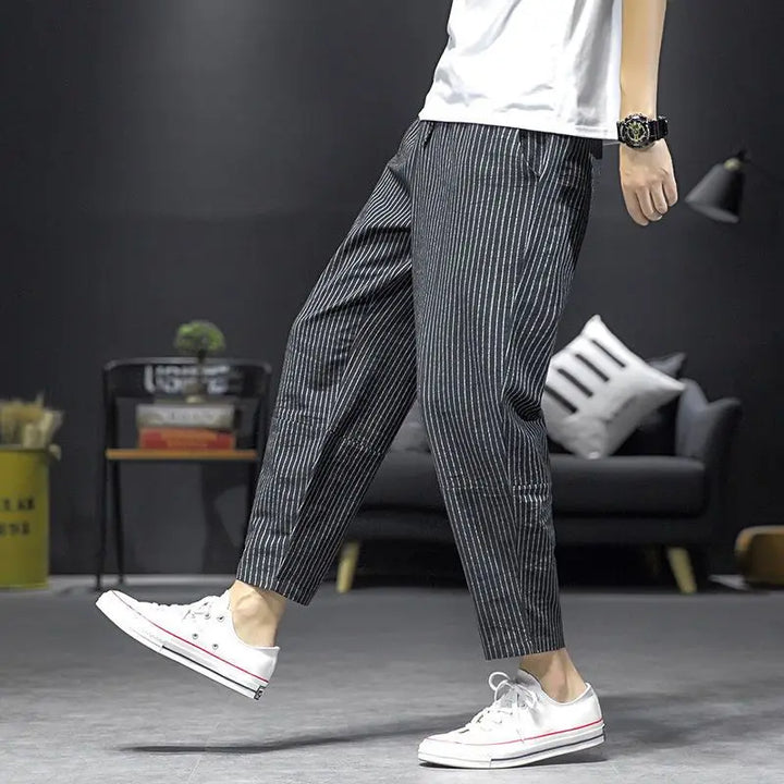 A person in a white T-shirt, casual straight leg pinstripe linen pants, and white sneakers walks indoors on a gray floor, with a black sofa and decor visible in the background.