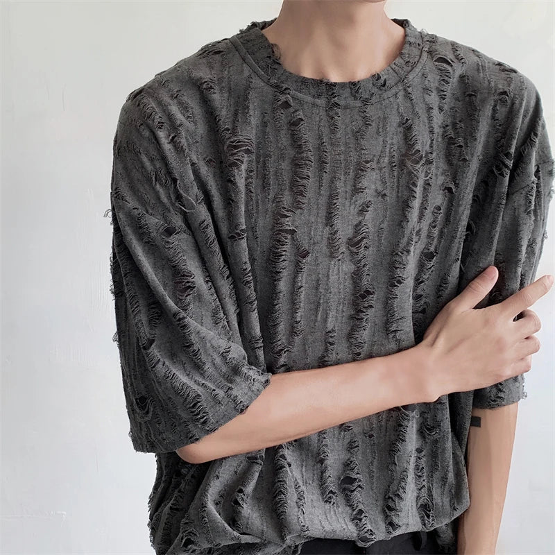 A person wearing a loose-fitting, dark gray, oversized tee made of distressed torn fabric with many rips and frayed edges stands against a plain light-colored background. Their left arm crosses their body, partially touching their right arm.