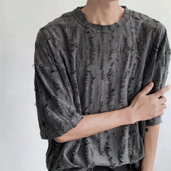 A person wearing a loose-fitting, dark gray, oversized tee made of distressed torn fabric with many rips and frayed edges stands against a plain light-colored background. Their left arm crosses their body, partially touching their right arm.