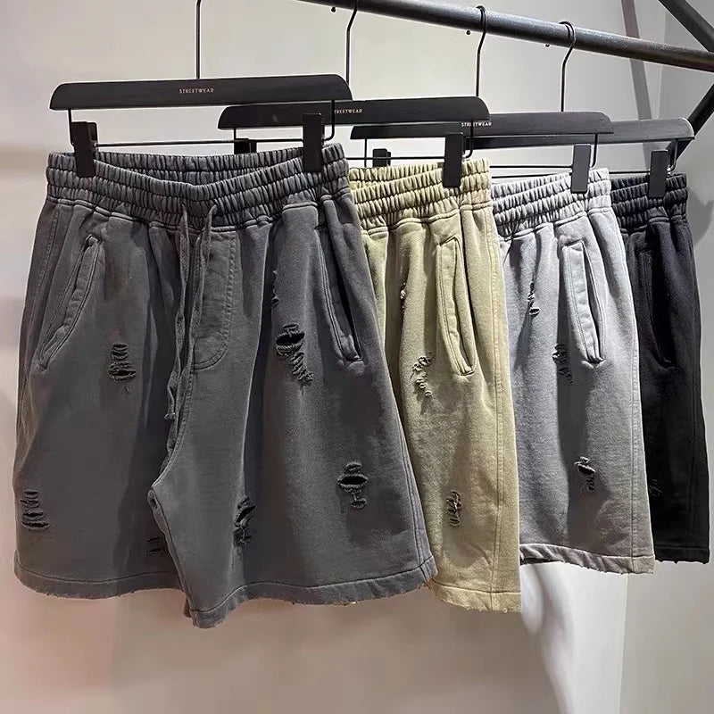 Four pairs of vintage baggy shorts in gray, beige, light gray, and black hang on black hangers from a clothing rack, each featuring an elastic waistband and a distressed look with ripped detailing.