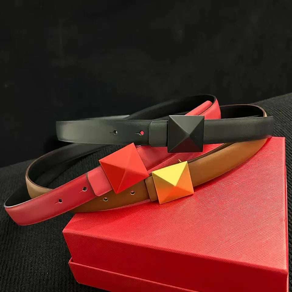 Three belts with large pyramid-shaped buckles rest on a red box. The top leather belt is black with a black buckle, the middle is reddish-pink with a red buckle, and the bottom is brown with an adjustable length and a gold buckle. The background is black.