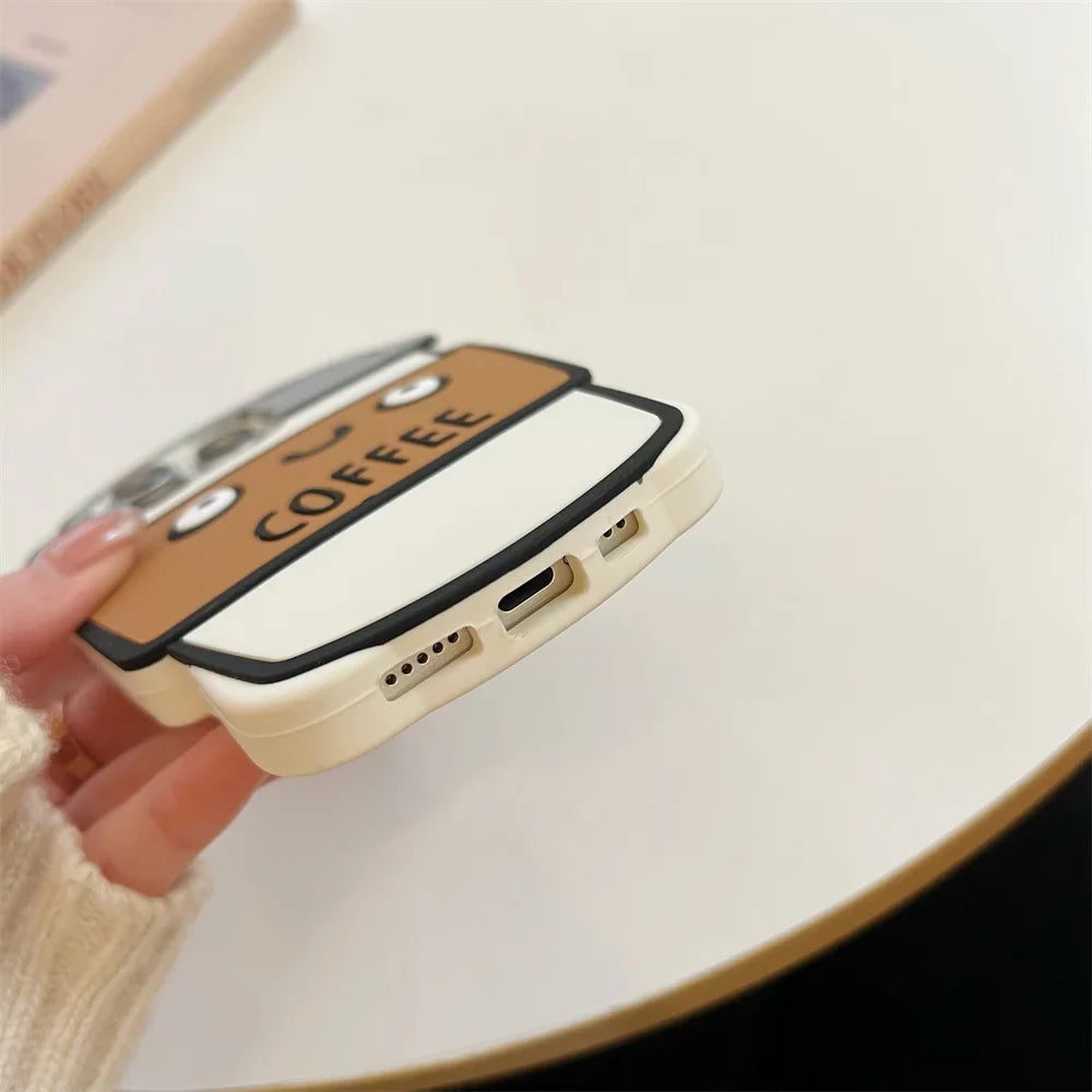 A hand holds a phone with a cute, cartoon-style 3D coffee cup case on a white tabletop. Crafted from soft silicone, the case reads COFFEE, features an illustrated face, and offers shockproof protection.