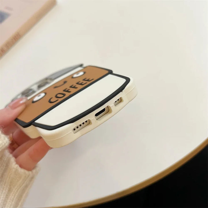 A hand holds a phone with a cute, cartoon-style 3D coffee cup case on a white tabletop. Crafted from soft silicone, the case reads COFFEE, features an illustrated face, and offers shockproof protection.