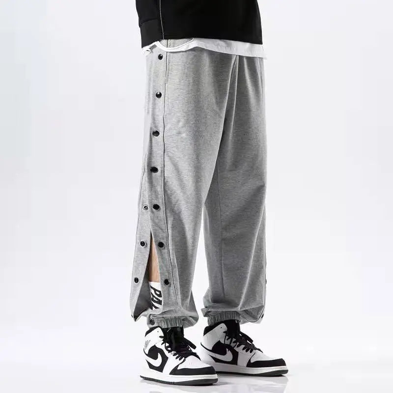 A person wearing gray button-up pants with black and white sneakers. The gym pants have side buttons partially undone, revealing some skin. Only the lower half of the body is visible.