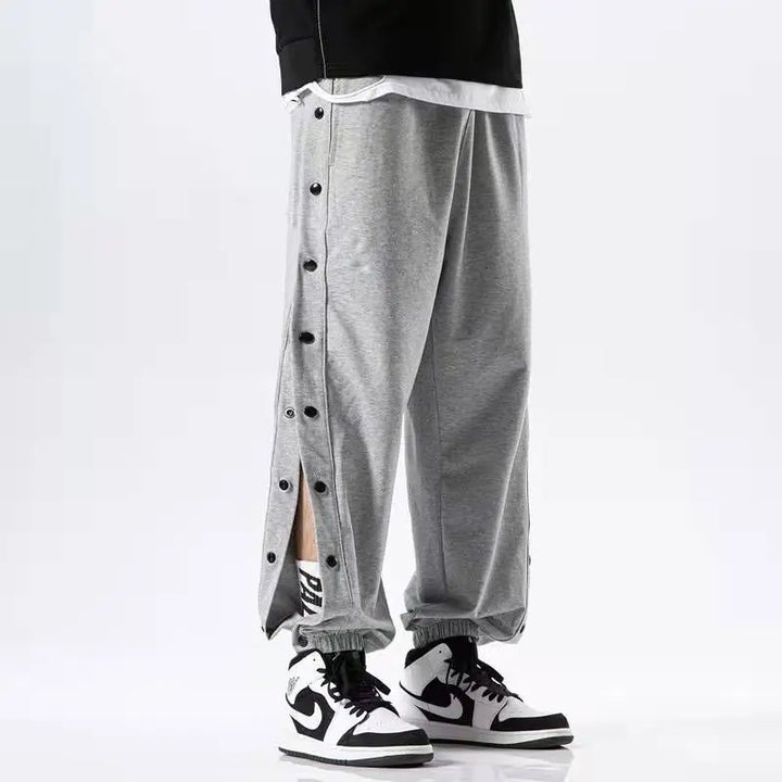 A person wearing gray button-up pants with black and white sneakers. The gym pants have side buttons partially undone, revealing some skin. Only the lower half of the body is visible.