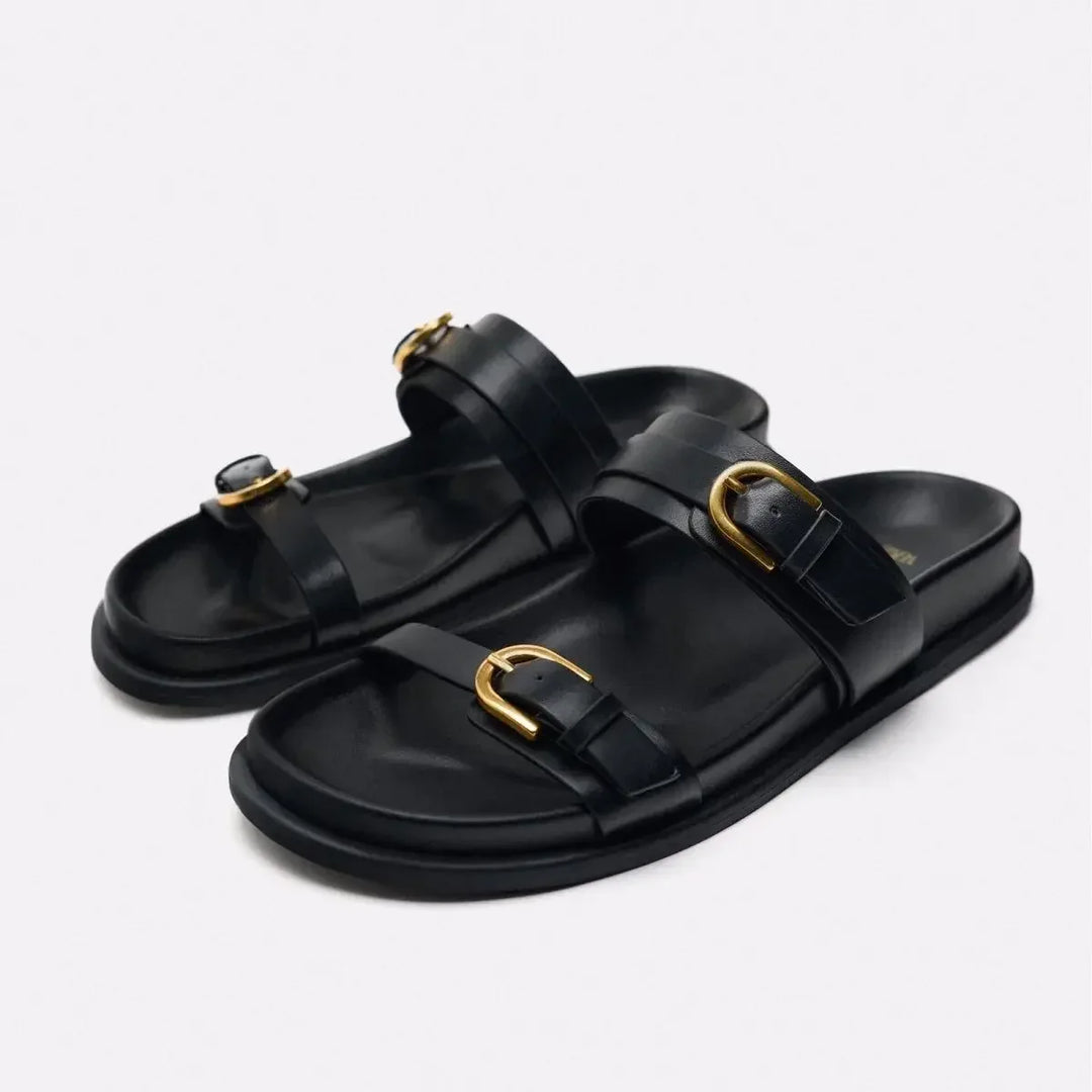 A pair of black slide sandals featuring a leather upper and two straps per sandal, each adorned with a gold buckle. The slip-on style offers effortless elegance, while the flat sole keeps the look simple and refined.