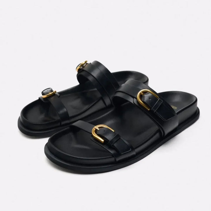 A pair of black slide sandals featuring a leather upper and two straps per sandal, each adorned with a gold buckle. The slip-on style offers effortless elegance, while the flat sole keeps the look simple and refined.