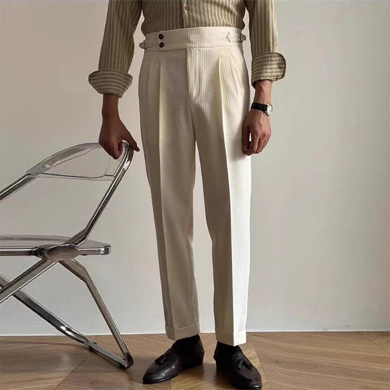 A person wearing luxury tailored pants, a beige striped shirt with rolled-up sleeves, black loafers, and a wristwatch stands beside a transparent chair on a wooden floor.