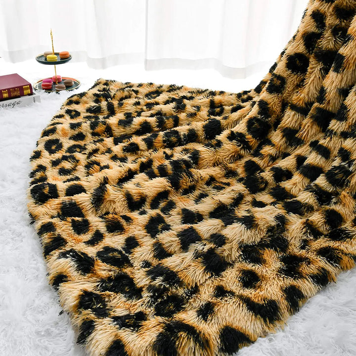 A plush throw with a black and tan leopard print blanket pattern is spread out on a white, shaggy rug. In the background, there are books and a tray with macarons near a window with sheer curtains.