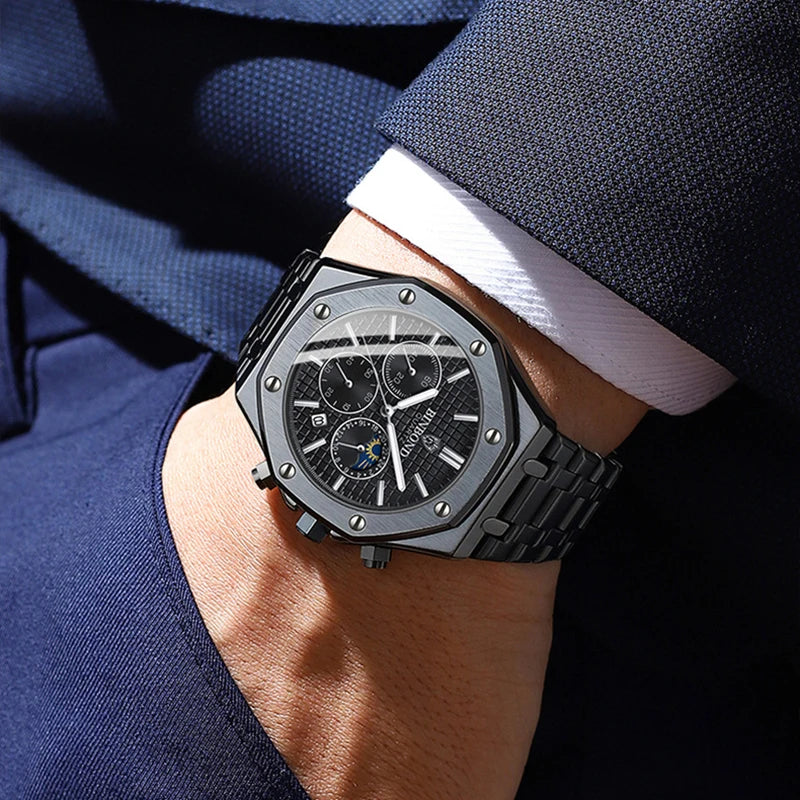 A close-up of a person wearing a dark metal stainless steel watch with a black dial, roman numerals, and chronograph dials, dressed in a blue textured suit with a white shirt cuff visible.