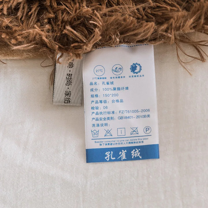 A close-up of two fabric care labels attached to a brown and white Soft Faux Fur Throw Blanket, with one label showing care instructions and product details in Chinese and icons for washing, drying, and ironing. Warm and Cozy Sherpa Blanket.
