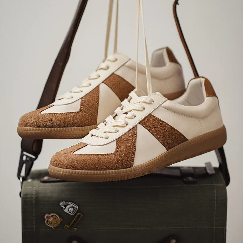 A pair of beige and brown lightweight breathable sneakers with premium cow suede and leather details hang by their laces over a green canvas bag decorated with small patches.