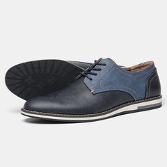 A pair of stylish mens lace-up patchwork oxfords in dark blue with denim-textured panels, white stitching, and a cushioned sole. One shoe is upright while the other is laid on its side to show the sole.